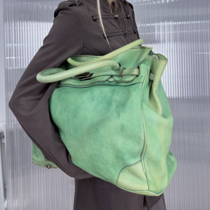 A person wearing a dark brown traditional-style jacket carries a large sage green leather tote bag with matching green handles and trim against a modern ribbed glass wall background.