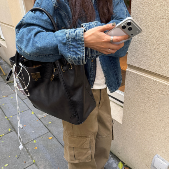 A person wearing a blue denim jacket and khaki cargo pants carries a large black leather shoulder bag with gold-tone hardware while holding a silver smartphone. White earphones are plugged into the bag, and the setting is an outdoor street corner next to a beige building.